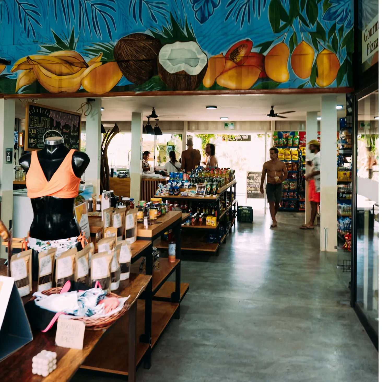 Interior de La Esquina con un mural de frutas tropicales al fondo, ropa colgada y estantes de producto.