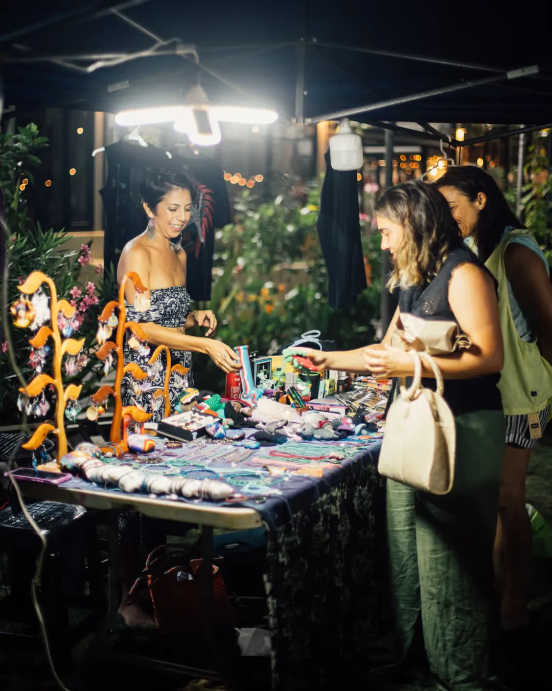 Puesto de artesanía en una noche de mercadito, iluminado con focos colgantes y gente mirando.