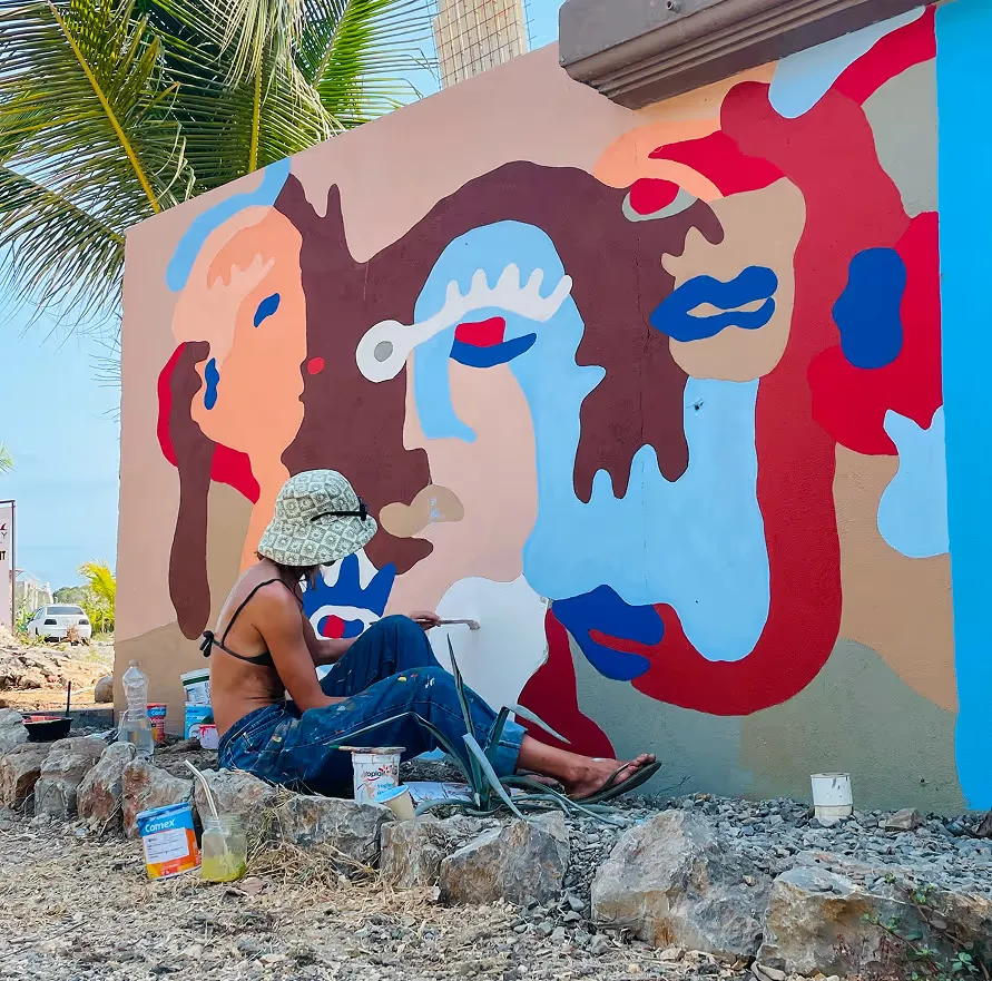 Un artista sentado sobre las rocas pintando un mural grande de rostros en rojo, azul y blanco.