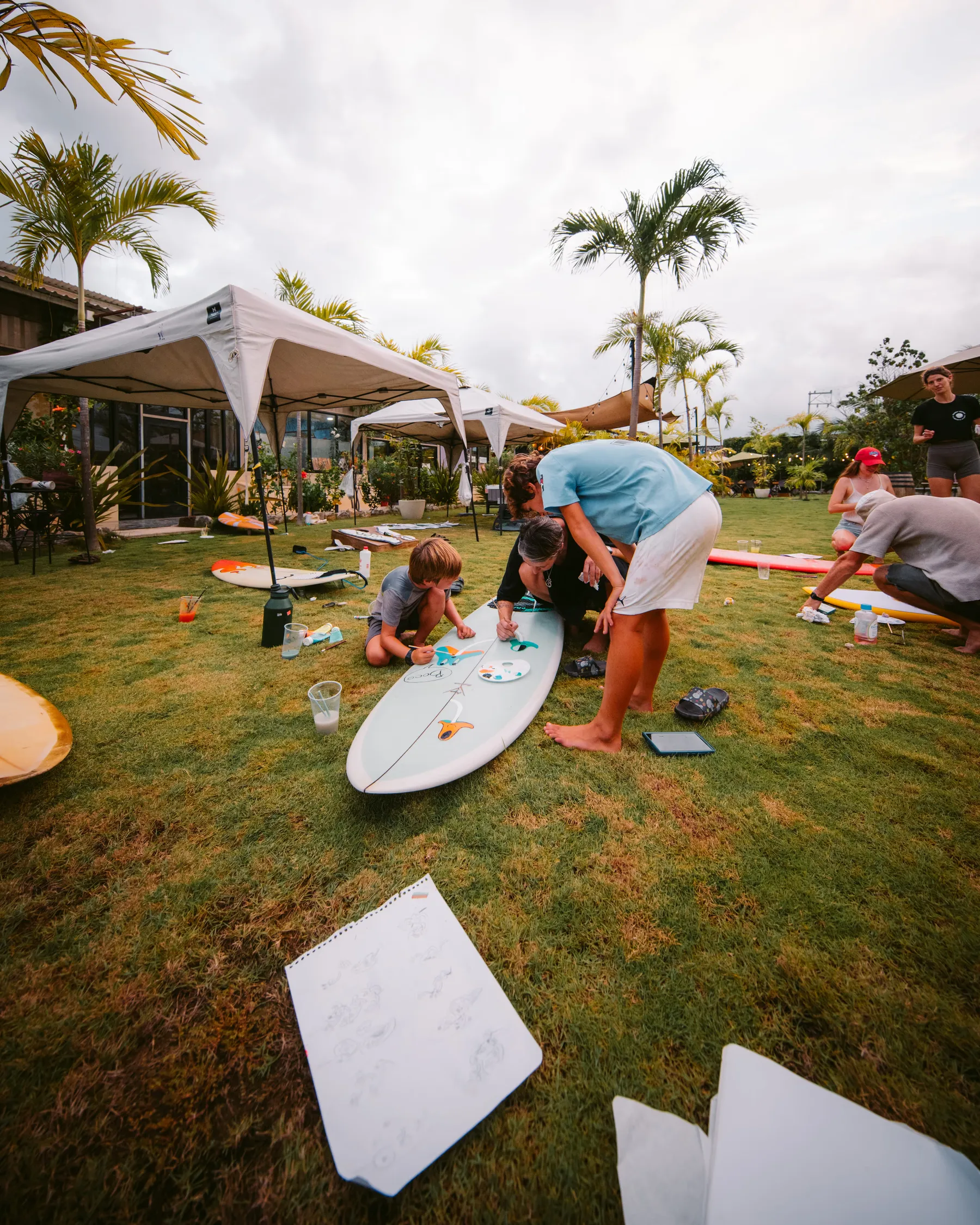 Niños pintando tablas de surf sobre el pasto, bajo una carpa blanca, con hojas de papel alrededor.
