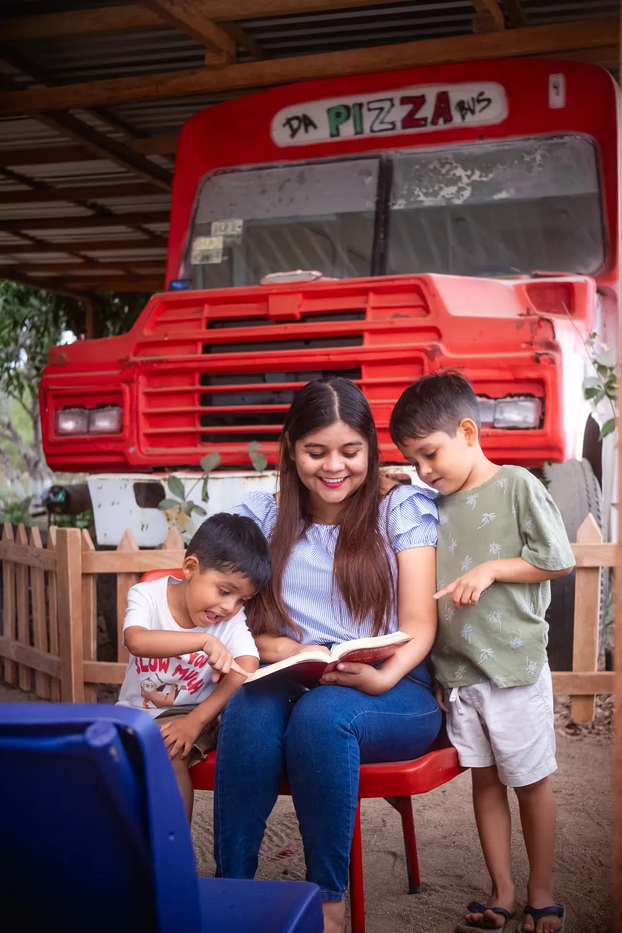 Camión de pizza rojo estacionado en el jardín, con una familia sentada afuera esperando.