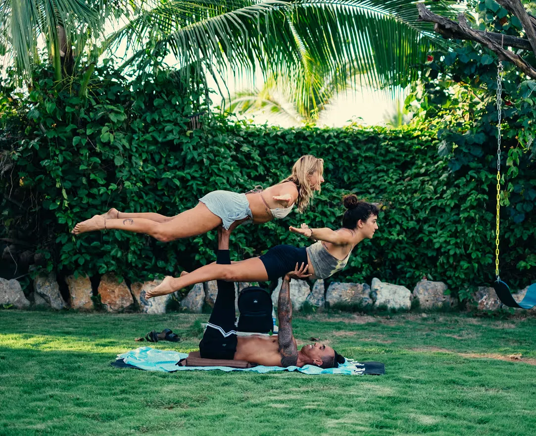 Clase de acroyoga sobre el pasto del jardín, frente a un seto verde y palmeras.